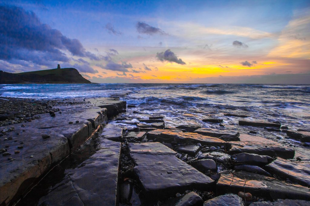 Rectangular rock stacks on the beach in Kimmeridge Bay around sunset with choppy sea beyond them and a low headland topped with a small round tower on the far side of the bay, with blue sky and a bright orange glow above the horizon