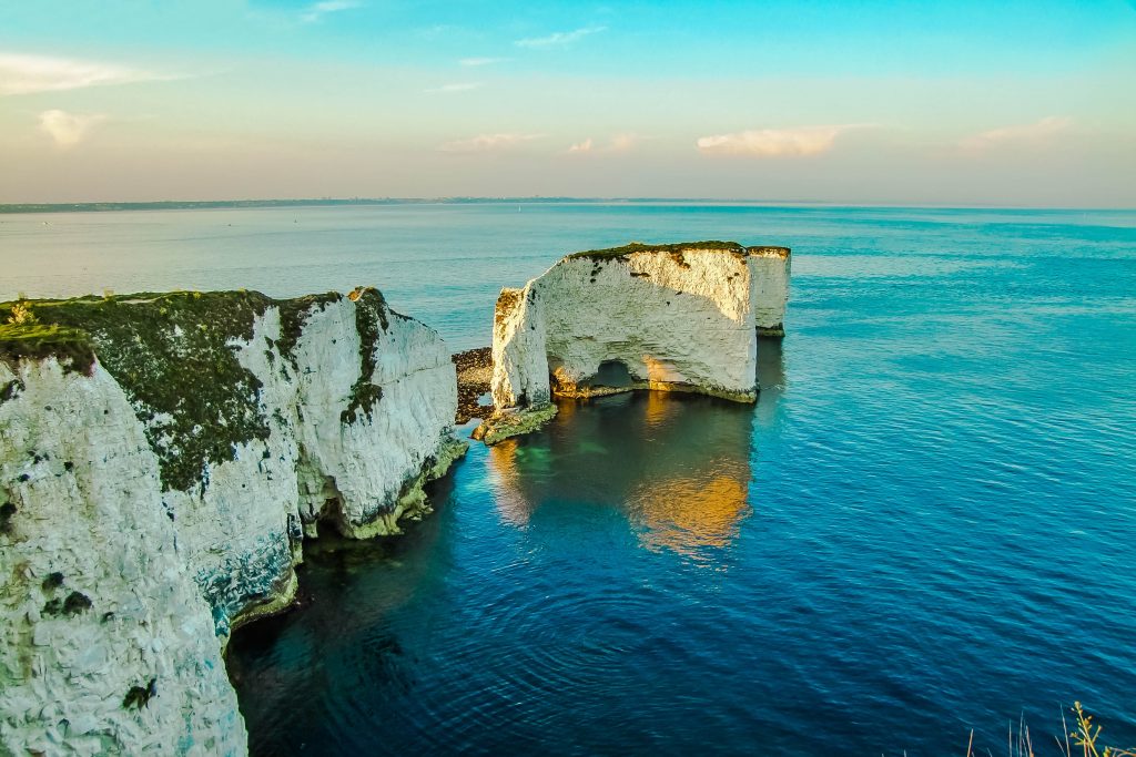 White cliffs with a few white rock stacks leading away from the end into the sea which is very blue and calm on a sunny day just before sunset with pink sky on the horizon - Old Harry Rocks in Dorset England