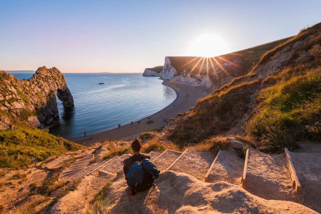 silhouetted woman sitting at the top of some stone and wooden steps leading down to a crescent shaped bay with a small beach and the rock arch of durdle door over the sea at the left side of the bay. Dorset Jurassic Coast highlights.