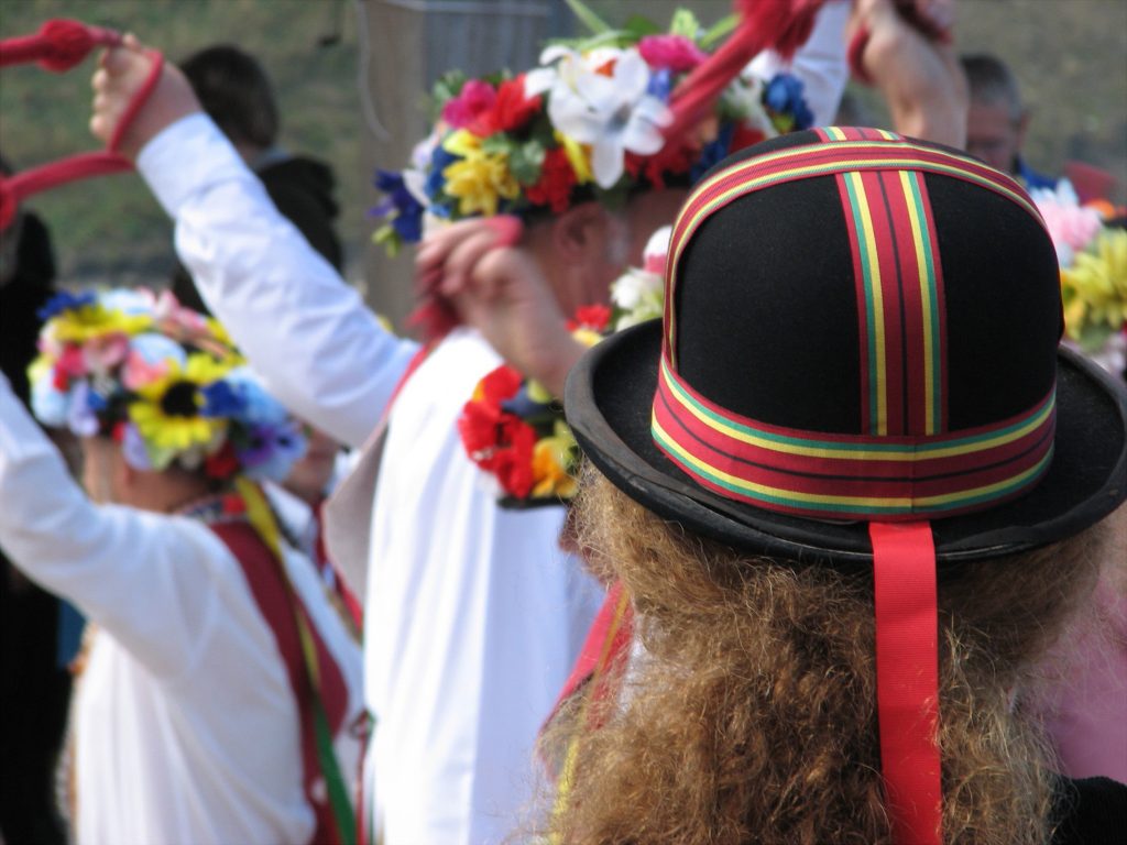 Close up of the back of a head with long curly brown hair wearing a black bowler hat with red green and yellow striped ribbon tied over it in a + shape, there are several morris dancers out of focus behind. Dorset Festivals and Events