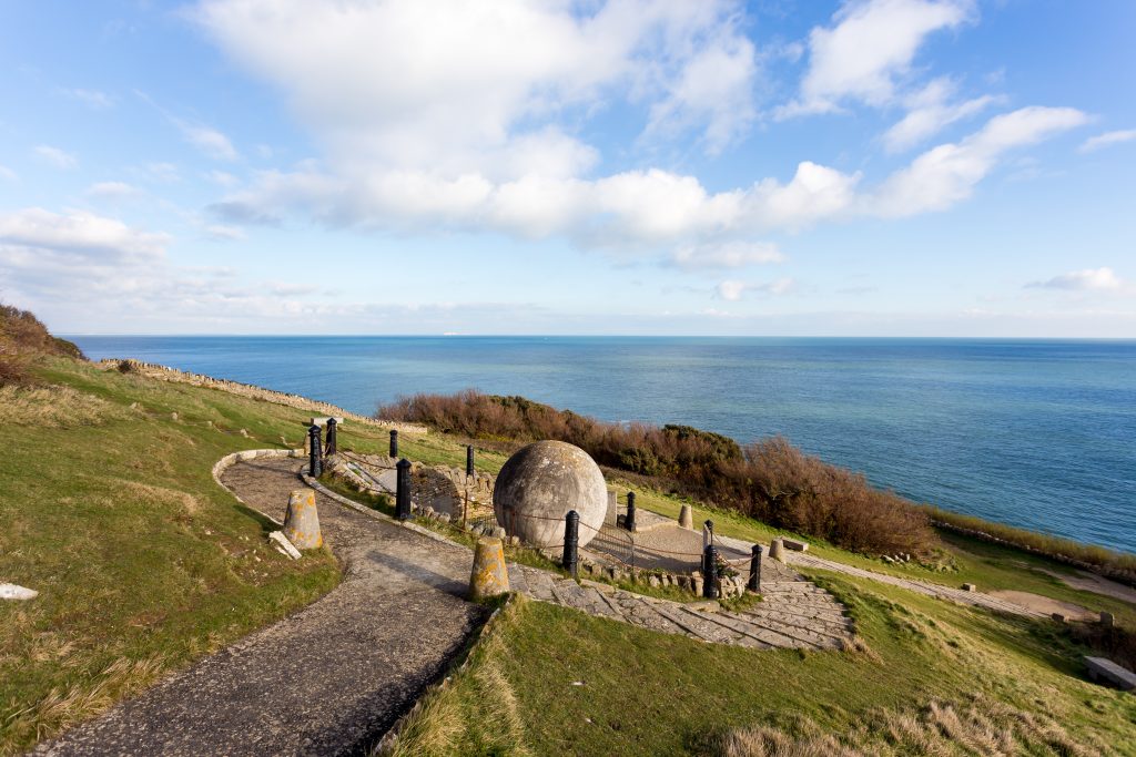 Large grey stone sperical carving of a globe on the side of a grassy sloping cliff with the blue sea behind and a stone pathway leading up to the globe which is encircled in metal railings. Durlston Country Park Dorset on a sunny day with blue sky. 