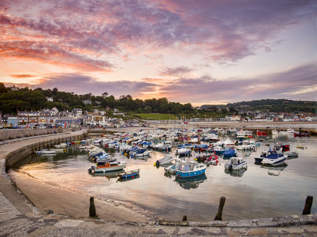 View of Lyme Regis Harbour looking back at the town from the harbour wall with a purple and pink sunset sky above and the harbour filled with small sailing boats. Things to do in Lyme Regis Dorset.