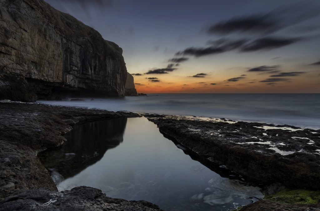 Very still calm rock pool on the flat rocks at Dancing Ledge in Langton Matravers taken just after sunset with a dark blue sky and an orange glow on the horizon over the sea and a low rocky cliff silhouetted against the sky to the left behind the rock pool