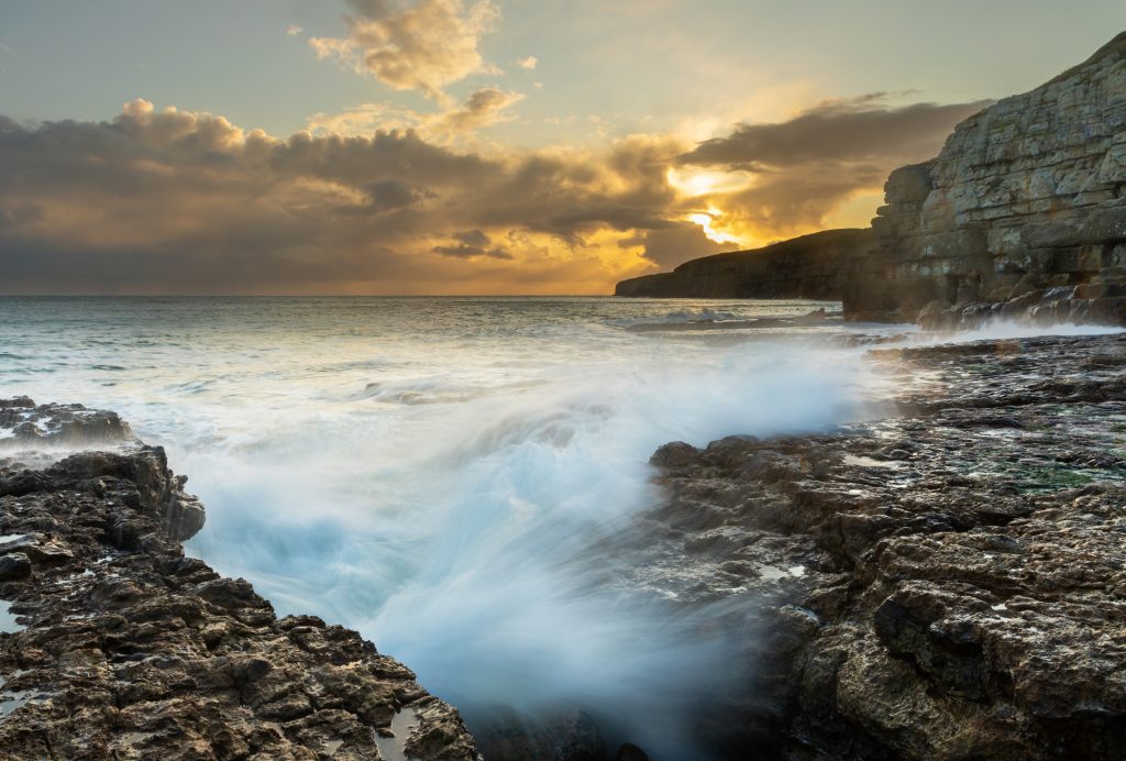 Small waves crashing in blurred motion against the rocks at Seacombe on the Dorset Coast with some low rocky cliffs in the distance. The sky and clouds are golden just after sunset. 