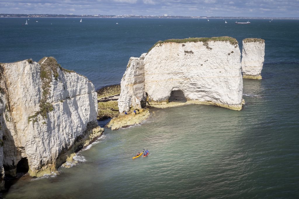 Two kayaks (one red one yellow) in the sea in front of the large chalk rock stacks called Old Harry Rocks with  the blue sea behind and the mainland far in the distance beyond. 