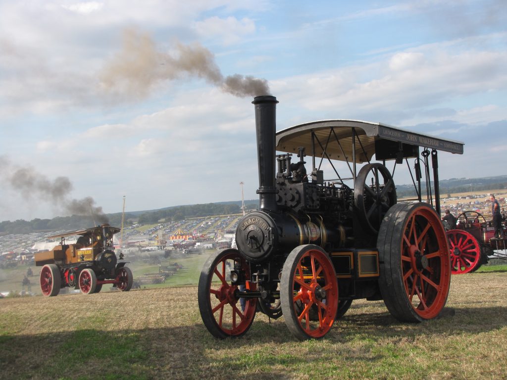 old fashioned steam tractor engine painted dark green with large red wheels in a grassy field at the Dorset Steam Fair with a large festival ground filled with stalls and crowds in the distance behind. 