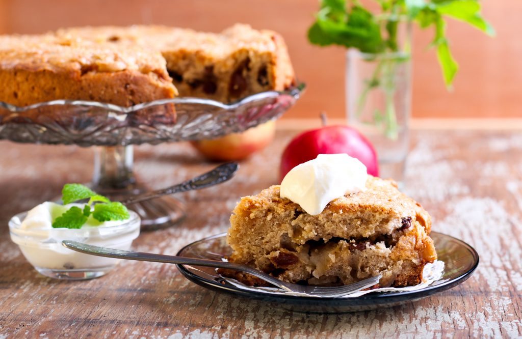 Close up of an apple cake on a grey plate topped with cream and the whole apple cake on a glass cake stand behind all laid out on a table with an orange wall behind. food and drink dorset england