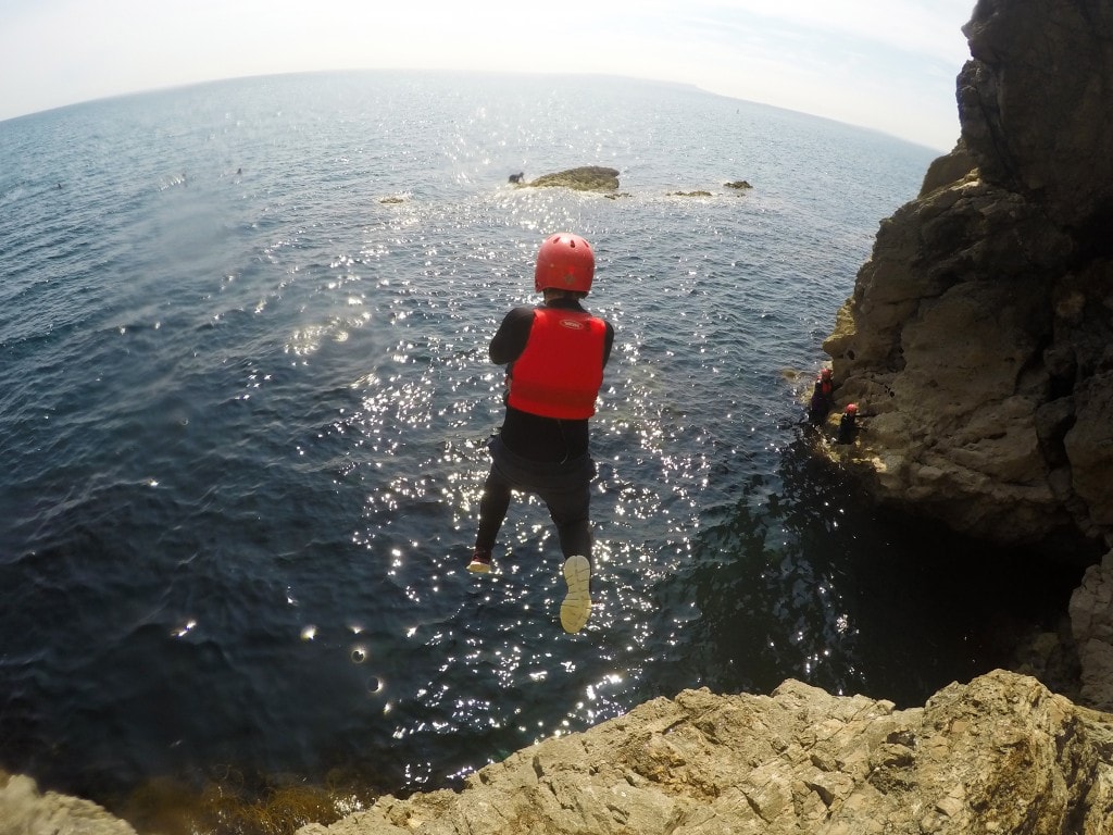 boy wearing a black wetsuit red life jacket and red helmet jumping intot he blue sea from a small cliff on a very sunny day while coasteering at lulworth cove