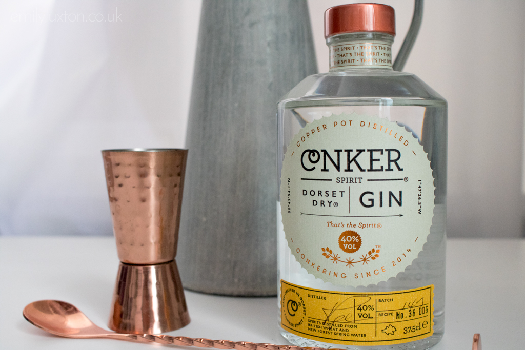 close up of a small glass bottle of gin with a copper lid and a circular white label printed with the name Conker Gin next to a small copper shot measurer and long cocktail stirrer