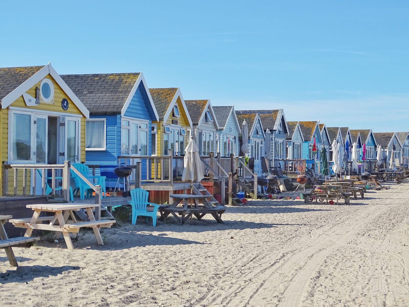 A row of beach huts painted alternatively yellow and blue with wooden picnic tables in front and a white sandy beach in front of that on a very sunny day with clear blue sky above - Mudeford Sandbank at Hengistbury Head in Christchurch Dorset England