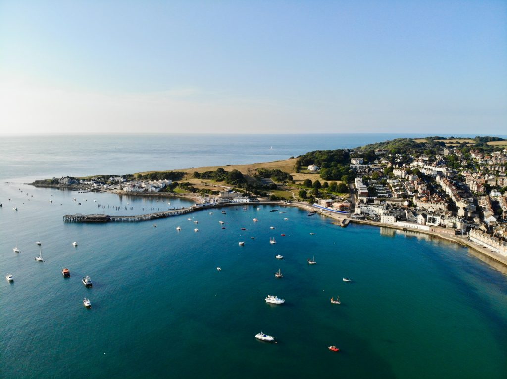 Aerial shot of Swanage Bay on a sunny day with the headland stretching out into the blue sea on the far side of the bay. there are many small boats moored in the bay and a small town along the shore. 