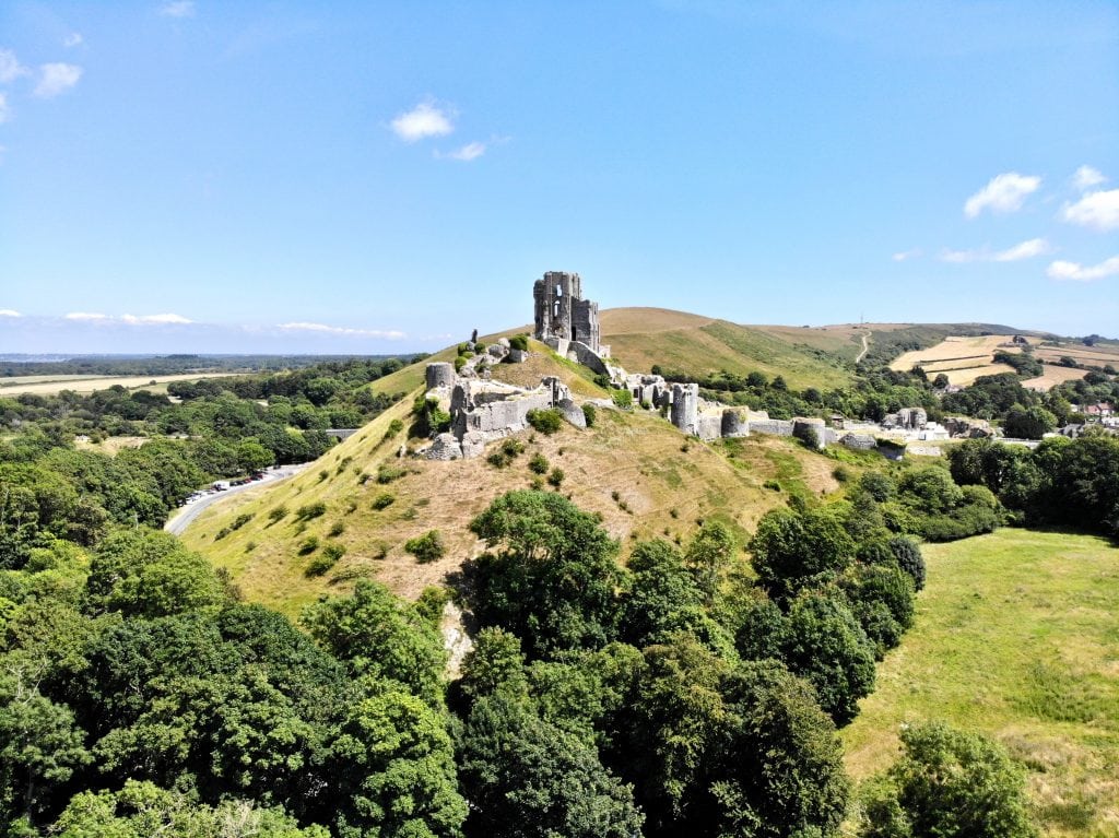 Ruins of a grey stone castle at the top of a grassy hill surrounded by woodland on a sunny day with blue sky above - Corfe Caste near Swanage