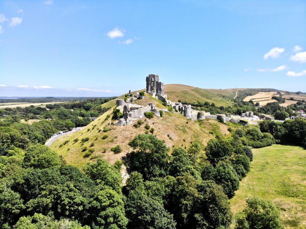Corfe Castle on a very sunny day - a small grassy hill surrounded by a green woodland with a ruined grey stone castle on top and blue sky overhead and a small village just visible in the distance behind the castle. Dorset England