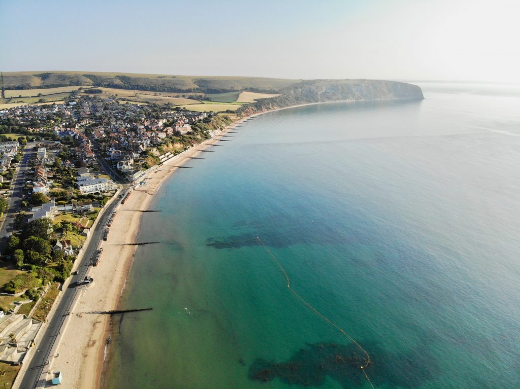 Aerial view of Swanage Beach taken on a sunny day with very bright blue and turquoise sea water and a long stretch of golden sand between the shore and the small town, with a grassy headland on the far side of the bay
