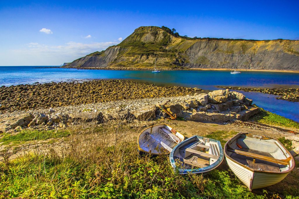 Three small wooden rowing boats on a patch of grass beside a shingle beach in Chapmans Pool on the Dorset Jurassic Coast with a rocky headland on the far side of the bay, taken on a very sunny day with blue sky above