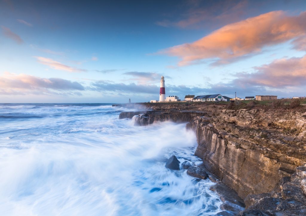Portland Bill at sunset with white capped waves crashing against the low brown rocky cliffs and a small white and red lighthouse in the distance just before sunset with pink clouds in the blue sky