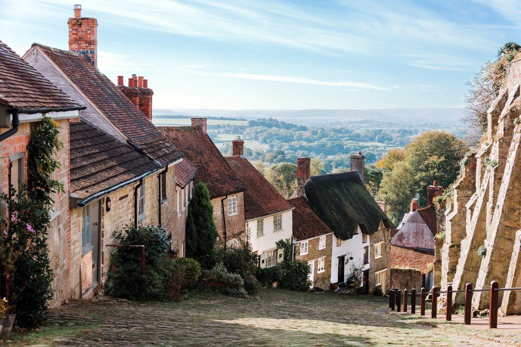 looking down a hill with grey flagstone road lined with small cottages with slate tiled or thatched roofs on the left and a brown stone wall on the right with green countryside visible in the distance - gold hill in Shaftesbury Dorset England 