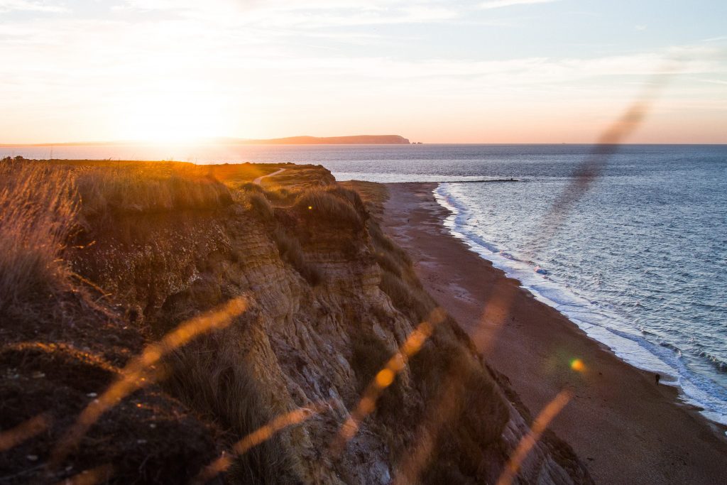 View of sunset from Hengistbury Head near Bournemouth - looking through some out of focus long grass and down a beige coloured sandstone cliff towards the beach. across the sea the sun is setting behing a silhouetted island. 
