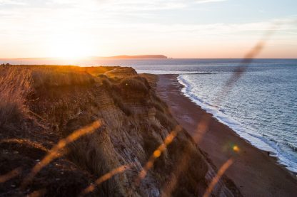 sunrise over a low headland with long grass out of focus in the foreground and the blue sea to the right