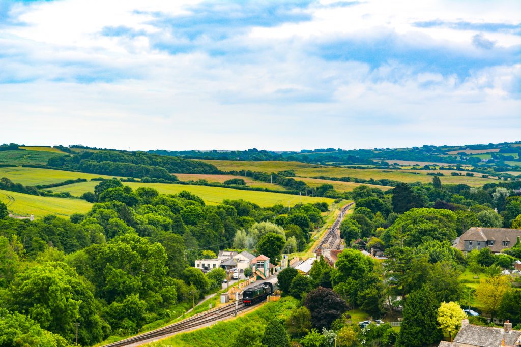 Aerial view of a black steam train outside a small old fashioned station with yellow signal box on Swanage Railway, with green countryside all around and a small woodland around the railway. 