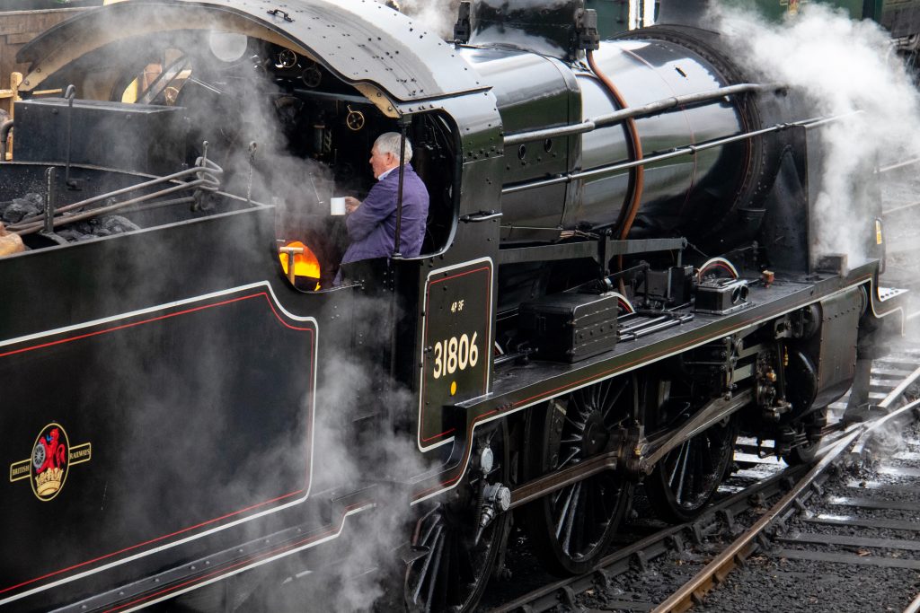 a man in the engine of a black steam train. the man is wearing purple overalls and has white hair and is holding a mug of tea. The train is surrounded by steam and has the number 31806 on the side. this is on the swanage railway one of the best things to do in dorset.