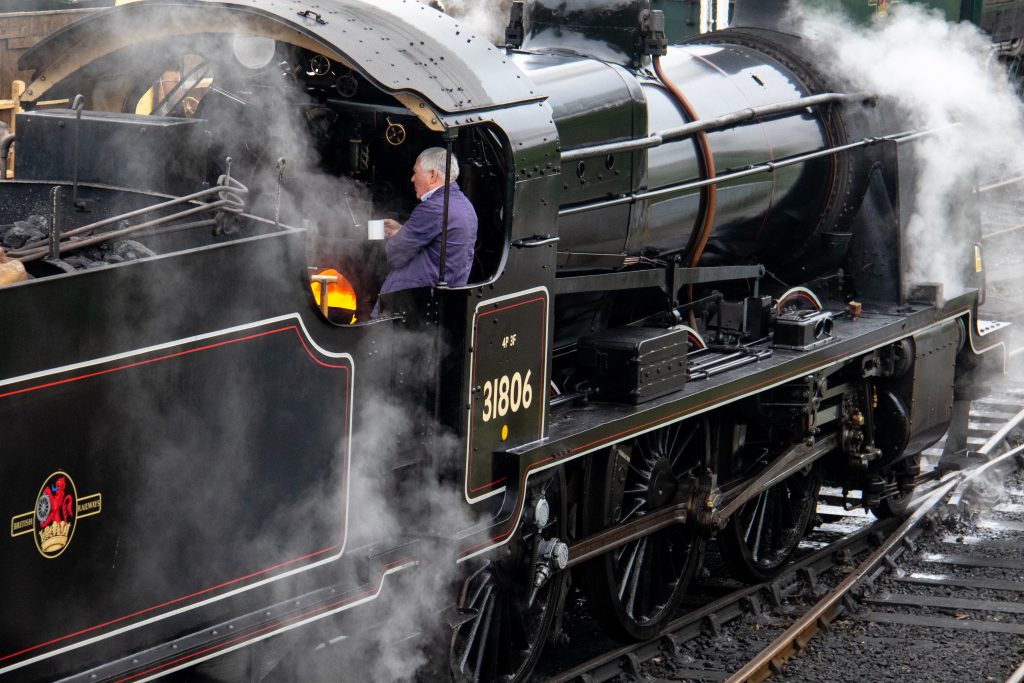 Exterior of a black steam engine with the number 31806 on the side and a grey haired man in blue overalls standing in the engine holding a white coffee mug. 