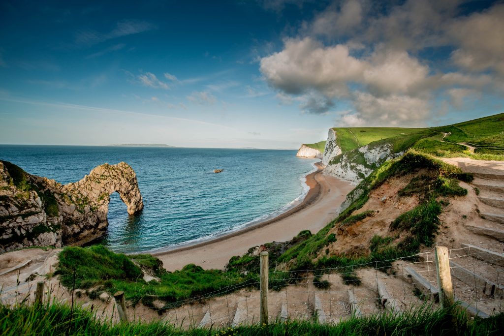 A set of stone and wood steps leading down towards a curved bay with a small shingle beach and the rock arch of Durdle Door over the sea at the left end on a sunny day with blue sea and blue sky overhead. How to get to Durdle Door