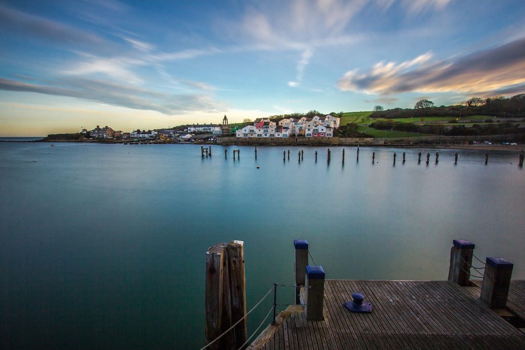 Looking across Swanage Bay at sunset with a small wooden jetty in the foreground and very still flat water in the bay. there is a grassy headland with a few houses on the far side of the bay and blue sky with golden clouds overhead. 