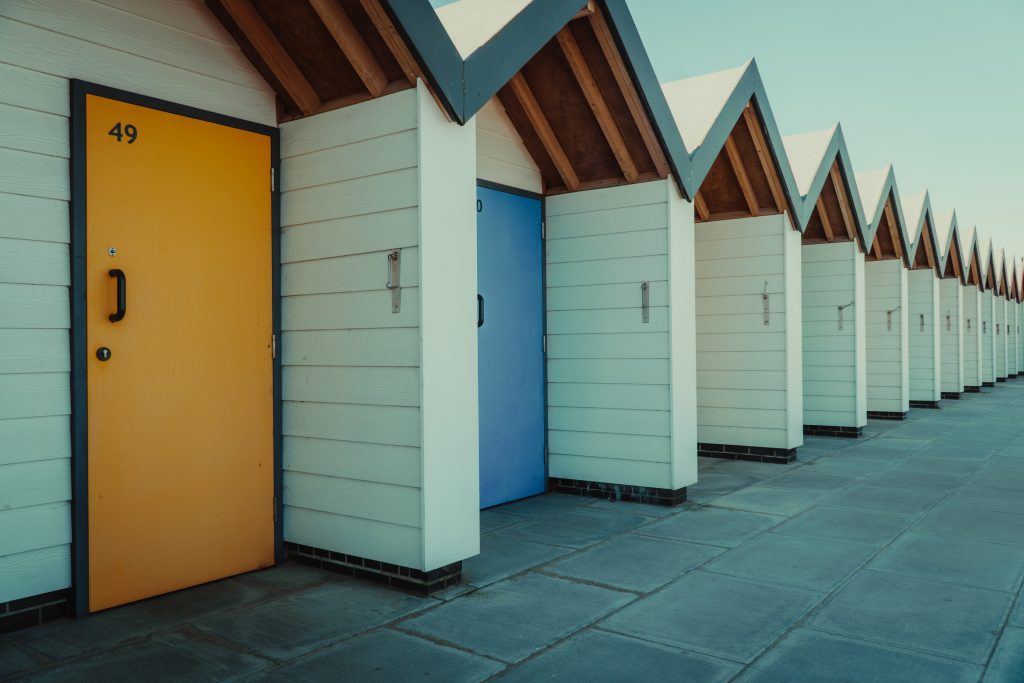 Row of white clapboard beach huts with blue traingular roofs, the closest has a yellow door and the next one has a blue door, with a paved walkway in front. 