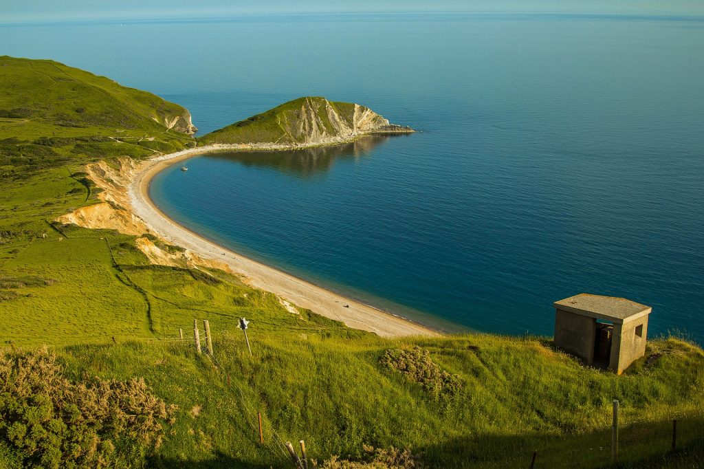 Looking down from a grassy clifftop at Worbarrow Bay on the Dorset Jurassic Coast with a long sandy beach around the bay and a small, narrow headland jutting out into the blue sea