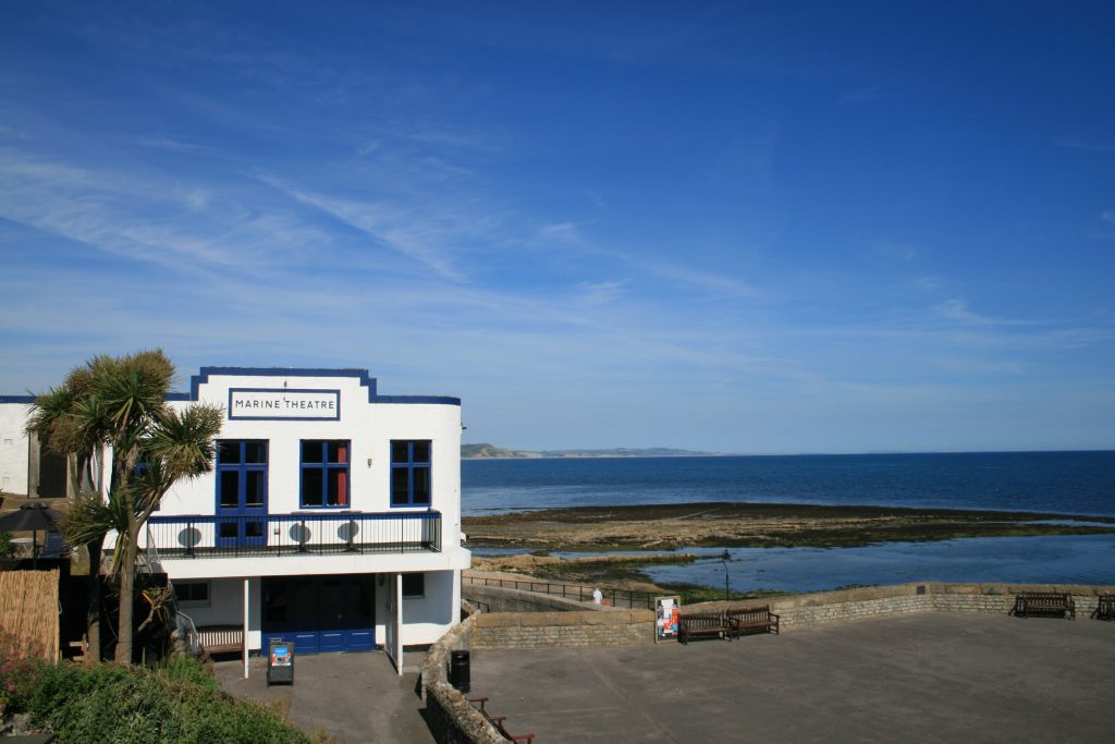 Exterior of the Marine Theatre in Lyme Regis next to the seafront with a bit of rocky beach and the blue sea to the right and some cliffs visible in the distance on a very sunny day with clear blue sky above. 