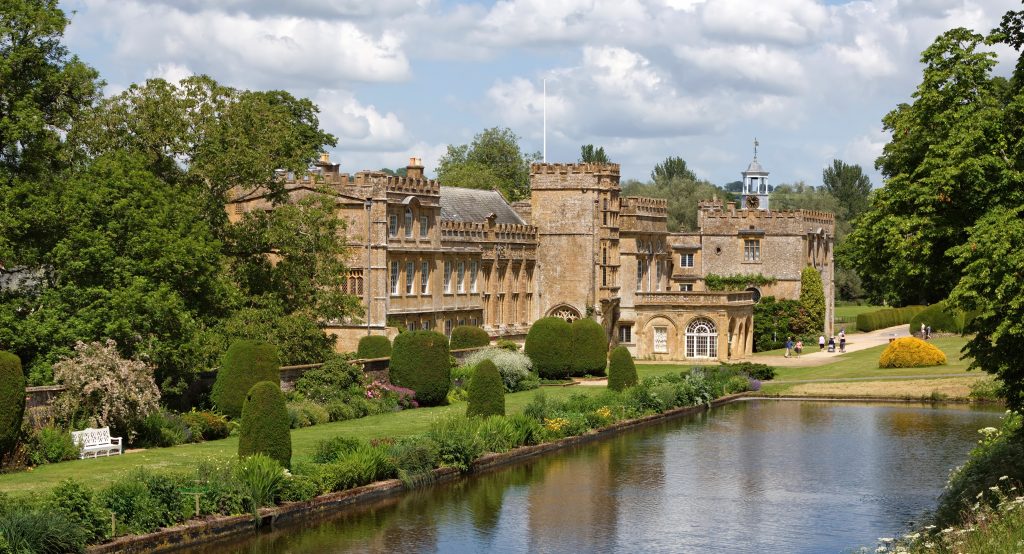 large stately home built from beige coloured stone with several turrets and a castle like appearance, surrounded by green leafy trees and a garden with small rounded hedges and a large rectangular pond in front. 