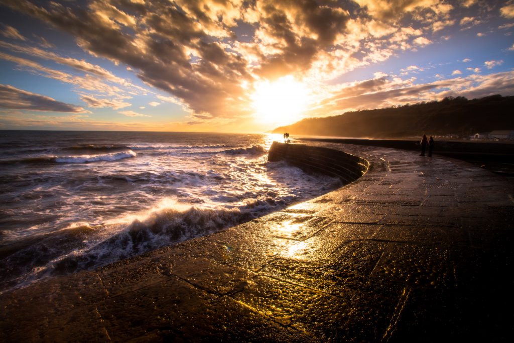 Low stone wall with a walkway on top next to the sea with waves crashing against the wall and a low headland in front silhouetted against the sky. The sun is setting over the sea and a couple walking along the wall are silhouttted in front of the sunset light. 