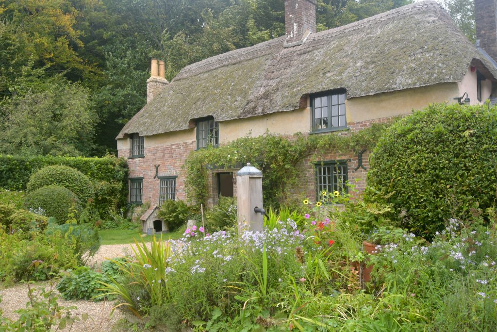 a two storey cottage bilt from red brick with grey window frames and a thatched roof with two chimenys. the cottage is in a garden filled with different colored flowers. 