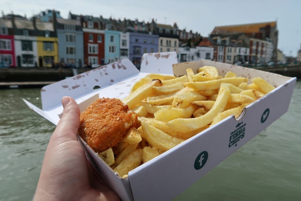 Hand holding a white cardboard box of fish and Chips in front of Weymouth harbour with colourful buildings on the far side of the water