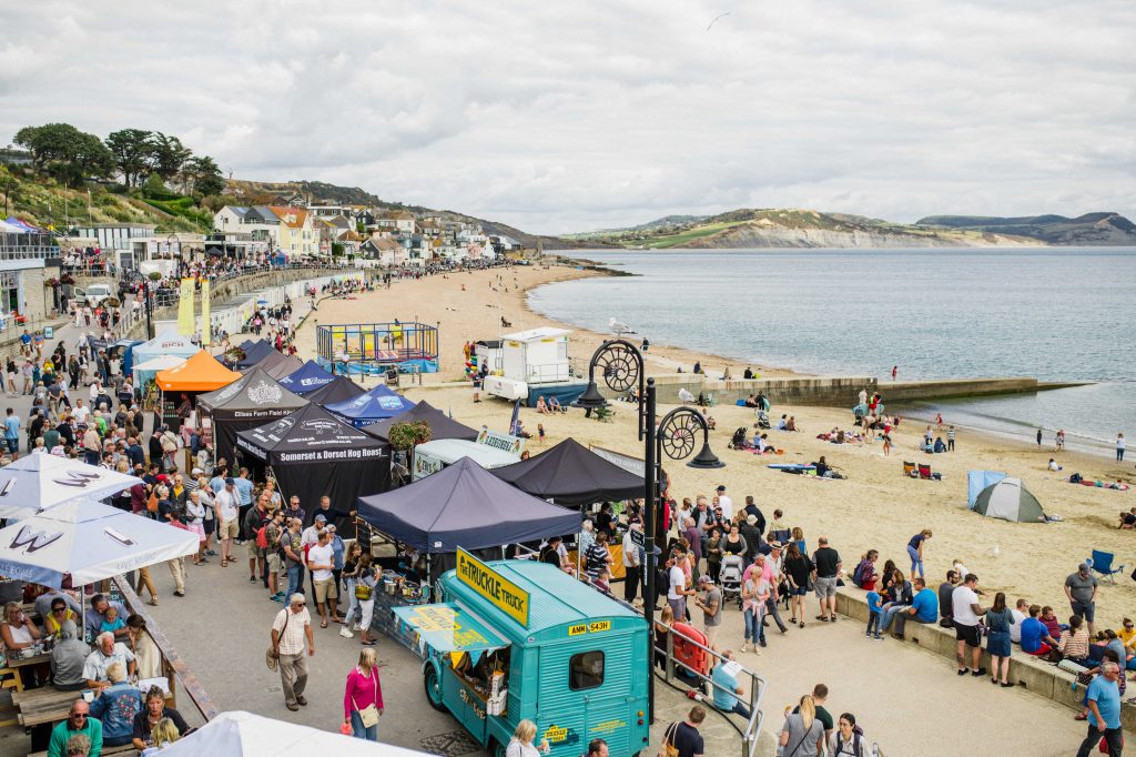 Food Rocks Festival in Lyme Regis, an aerial shot of the sandy beach with a low grassy headland visible across the bay in the distance, the paved esplanade next to the beach is filled with canvas food stalls and a bright turqouise food truck with a large crowd of people walking between them all. 