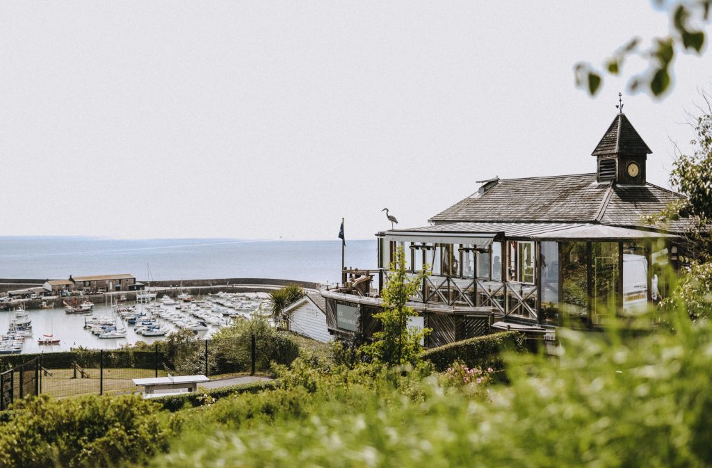 Hix Restaurant in Lyme Regis, a small wood and glass building with a grey tiled roof on the edge of a low hill above a harbour filled with small white boats with the sea behind that. there is a heron stood on the roof of the restaurant and a green leafy bush in the forgeround.