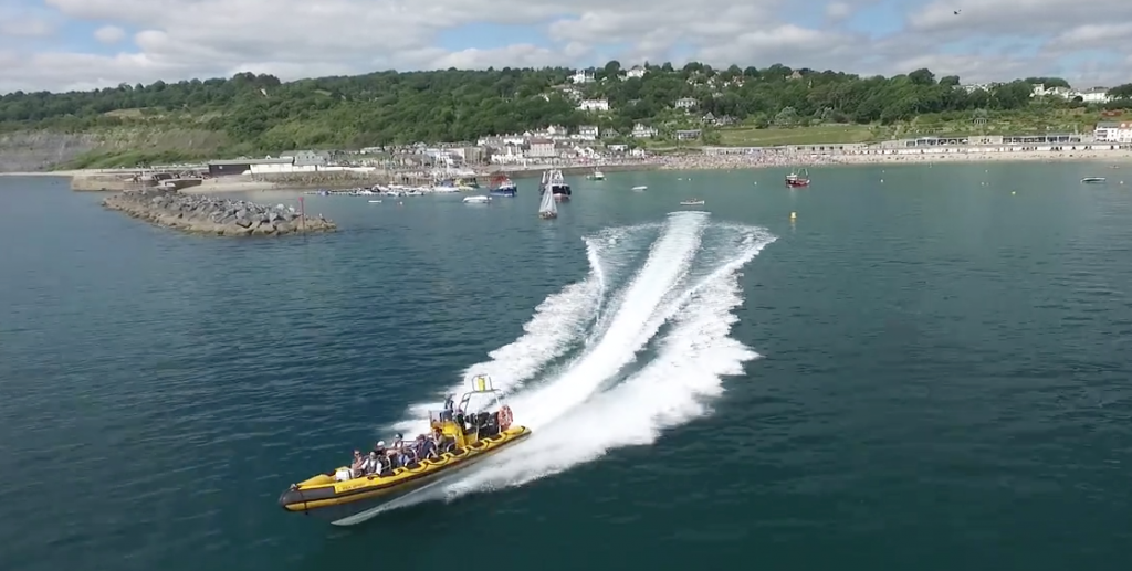 Aerial shot taken above the sea looking back at Lyme Regis with the stone breaker pointing into the sea and a few sail boats near the harbour entrance in the distance, there is a bright yellow and black RIB boat with several people on board going very fast through the water close to the camera with a white frothy boat trail behind it.