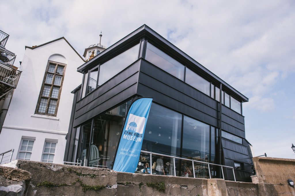 Exterior of Lyme Regis Museum, a modern two storey building made form black metal with large glass windows all around both floors, and an older whitewashed townhouse just behind it, shot from below with a cloudy blue sky overhead. 