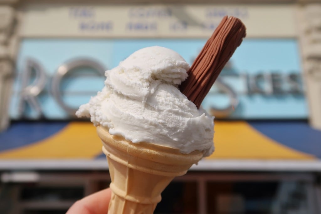 hand holding an ice cream cone with white ice cream and a flake in front of the blue sign for Rossi's Ices and the blue and yellow shop awning. Places to eat in weymouth dorset