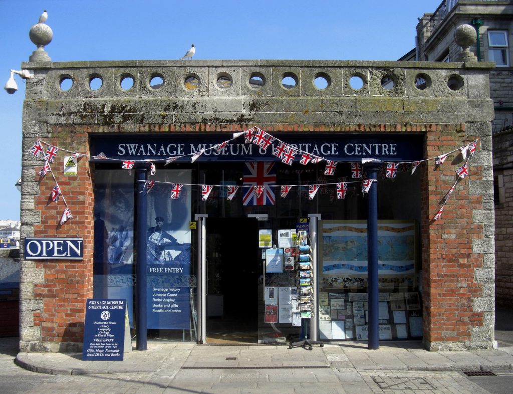 Exterior of Swanage museum, a one storey square shaped building made from red brick with a dark blue sign above the doorway, taken on a sunny day with blue sky above. 