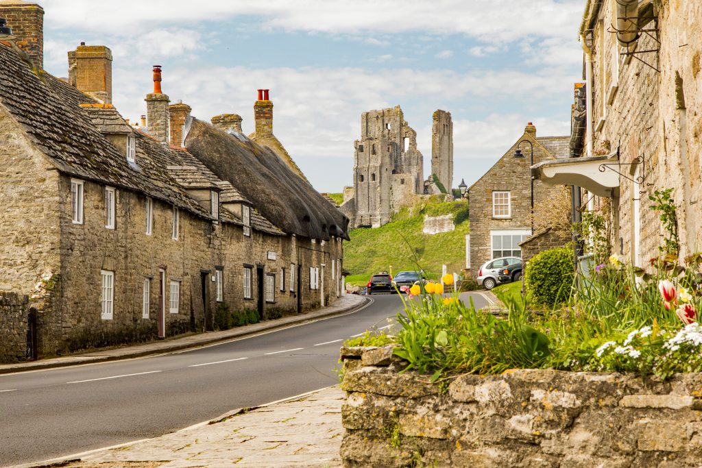 a road in a village with a row of terraced cottages wiht beige stone and grey slate tiled roofs and white framed windows. the furthest cottage has a thatched roof. At the end of the road is a grass hill with a ruined castle on top of it. Corfe Castle. 