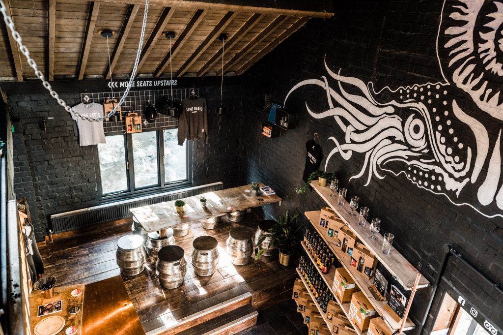 Interior of the Lyme Regis Brewery in Dorset, looking down from up high at the shop floor with black painted brick walls and a large white mural of a sea creature on one wall. there is a long wooden chair on the wood floor with chairs made from silver beer barrels and the walls are lined with wooden shelves stocked with beers. 