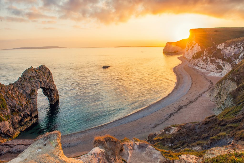 view of durdle door beach from the top of the cliff at sunset, there is a rock arch to the left and a curved shingle beach below the the cliffs and the sun setting over the sea taken on the walk from Lulworth Cove to Durdle Door.
