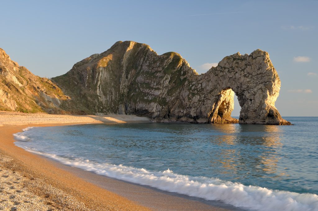 golden sand and shingle beach with calm sea and the large rock archway of Durdle Door over the sea taken around sunset with golden light and clear blue sky.