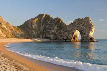 golden sand and shingle beach with calm sea and a large rock archway over the sea taken around sunset with golden light and clear blue sky. Lulworth Cove to Durdle Door Walk