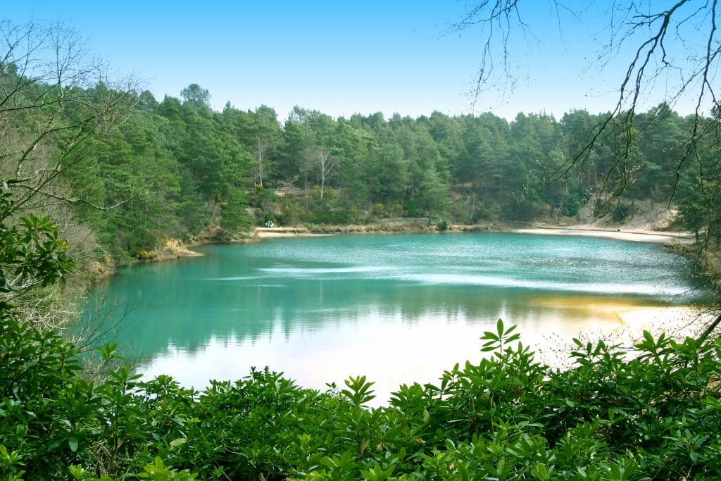 large pool with cyan water surrounded by pine trees with blue sky overhead. Blue Pool near Wareham on the Isle of Purbeck