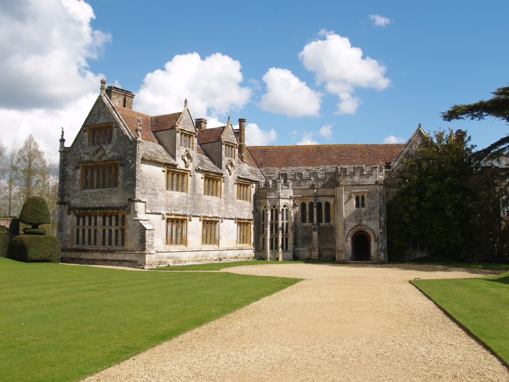 Athelhampton House - a large historic mansion made from grey stone with a red tiled roof and a very neat grassy lawn in front on a sunny day with blue sky above
