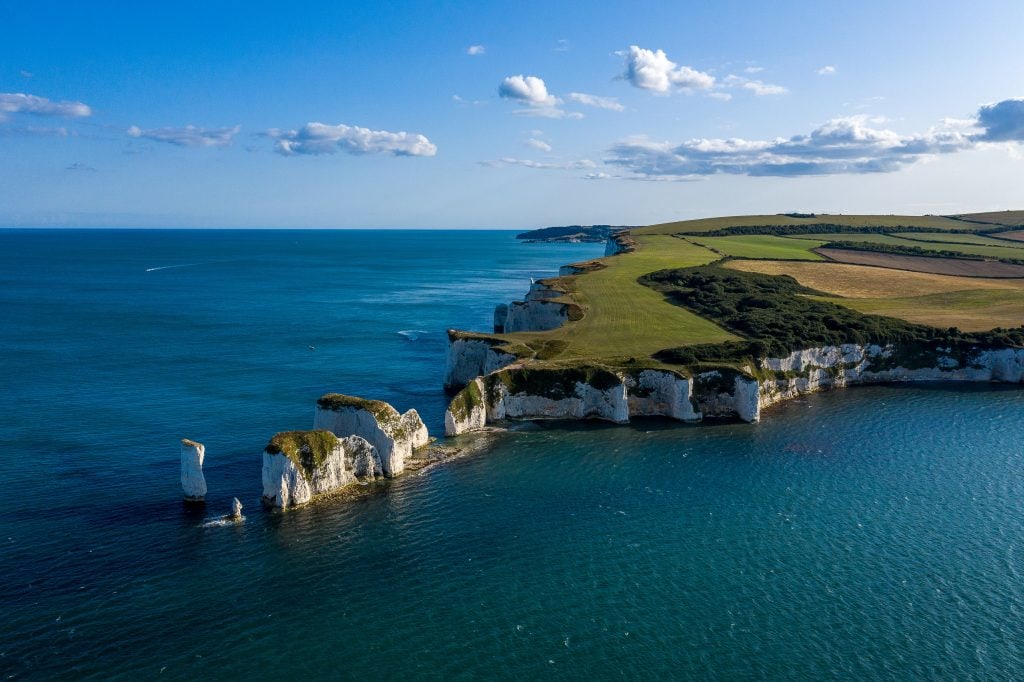 aerial shot of a grass topped headland with white cliffs surrounded by blue sea on a sunny day. A series of white chalk stacks extend out into the sea, some of them have a fuzz of green grass on top. The isle of purbeck is one of the best things to do near bournemouth. 