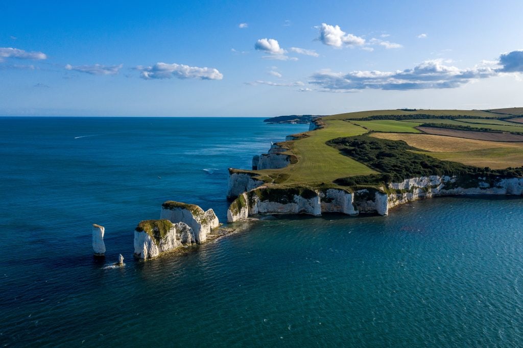 Arial shot of Old Harry Rocks on the Isle of Purbeck, a small peninsula of land covered in grassy fields with white chalk cliffs around the edge and a series of tall white rock stacks leading out into the blue sea